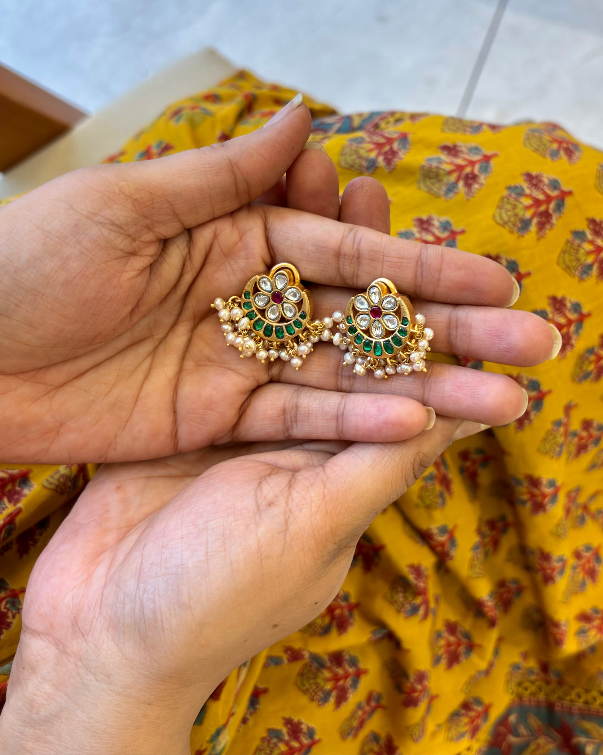 Gold earrings with green and red stones held between fingers against a yellow fabric background