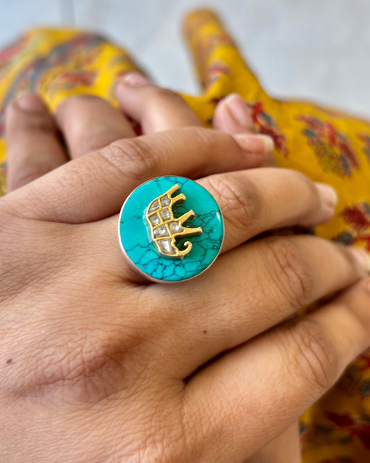 Turquoise ring with gold elephant design on a hand against a colorful fabric background