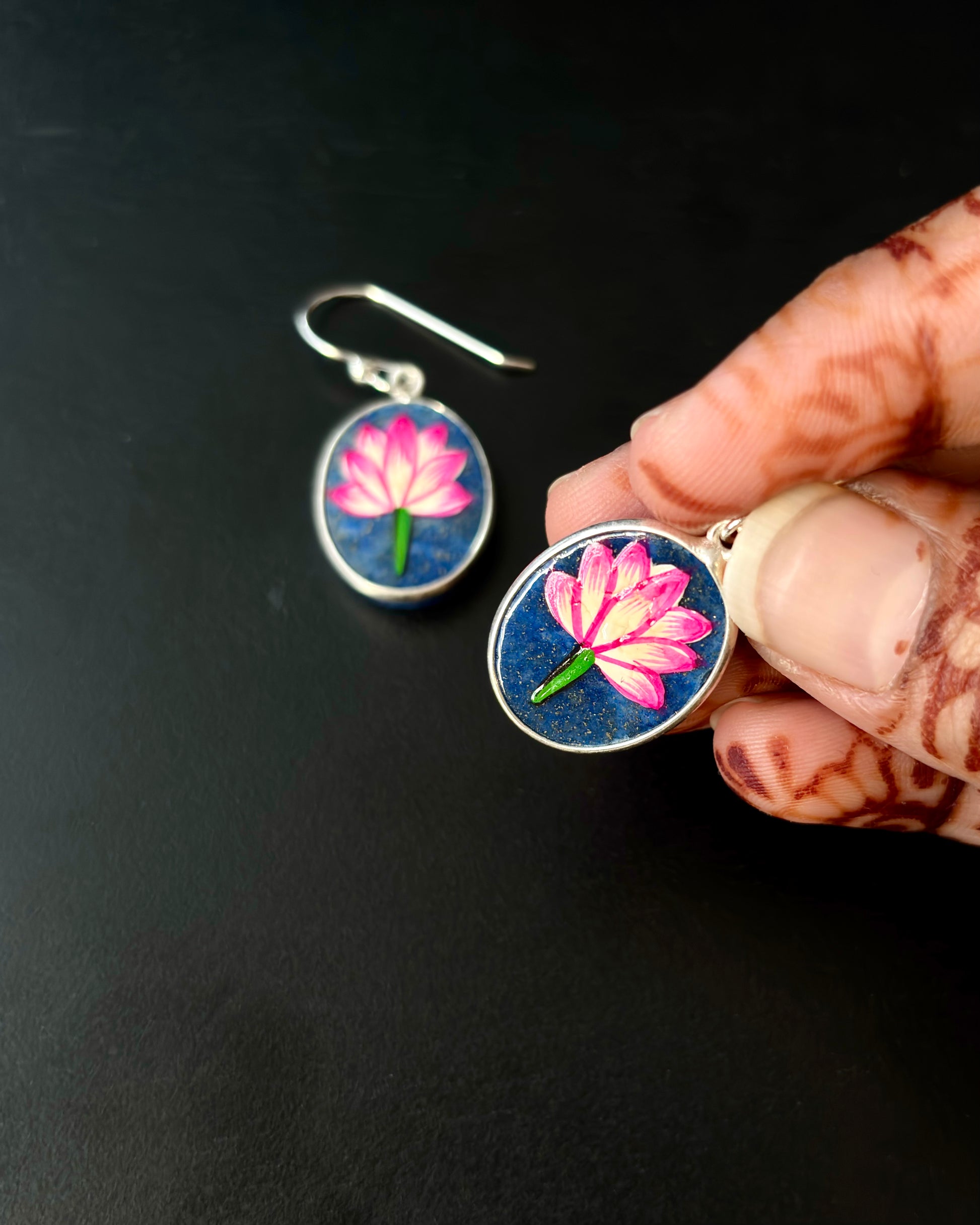 Hand holding a pair of earrings with pink flower design on a black background