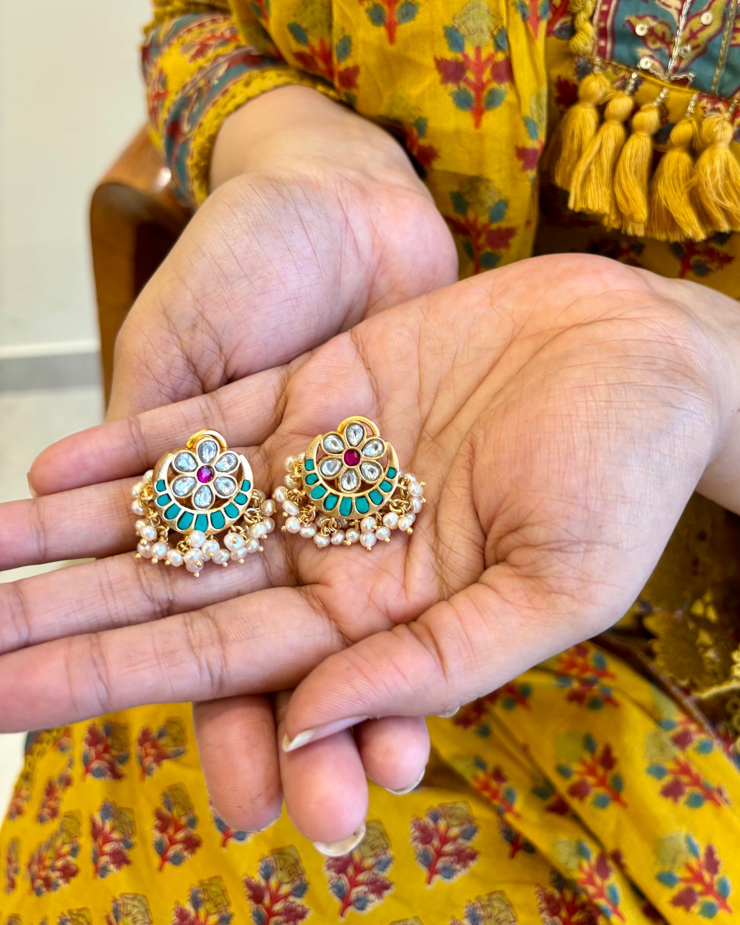 Pair of decorative earrings held between two hands with a colorful traditional outfit in the background.