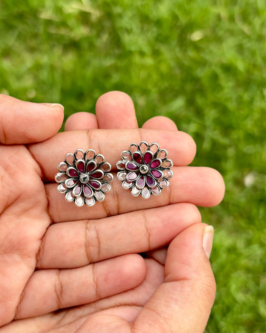 Floral silver and red earrings held between fingers against a green grass background
