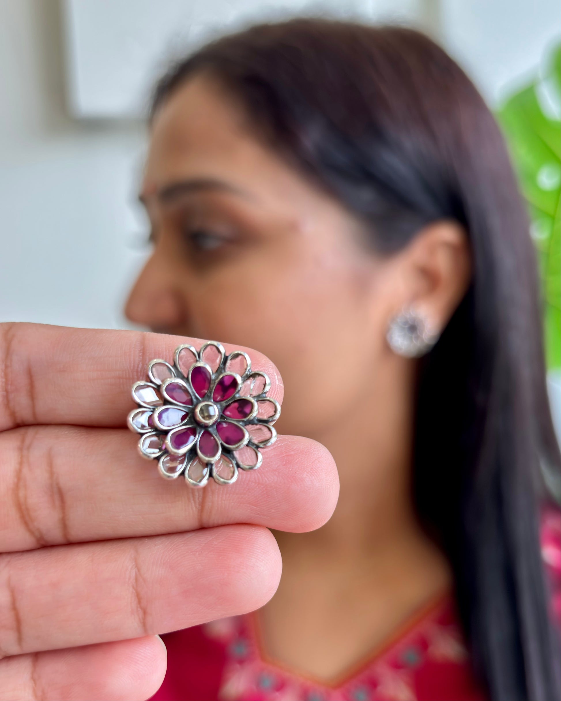 Close-up of a silver floral earring with red stones held by a hand, with a blurred background of a person wearing another earring.