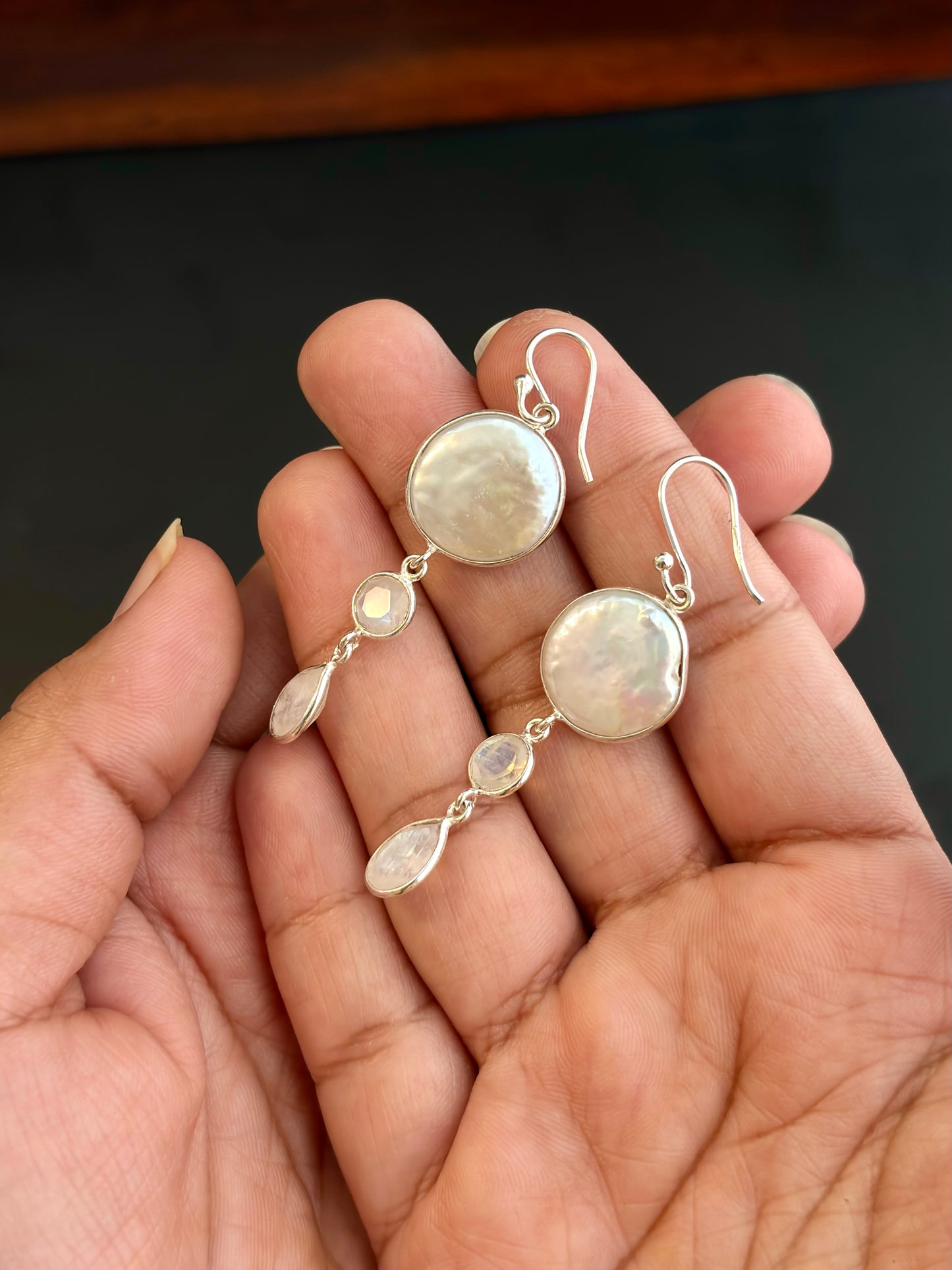 Silver earrings with round and teardrop-shaped gemstones held in a hand against a dark background