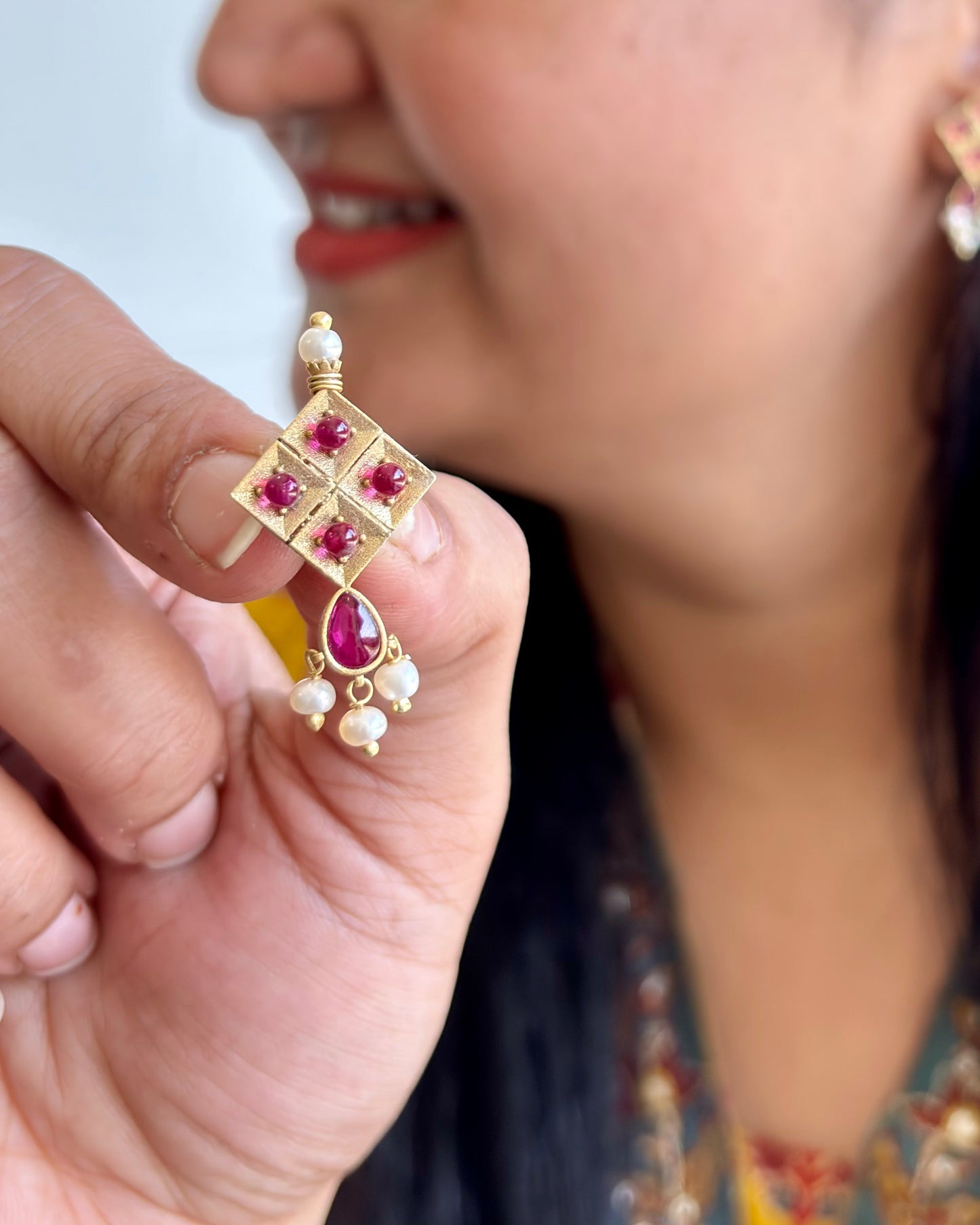 Gold earring with red gemstones held by a hand, blurred background