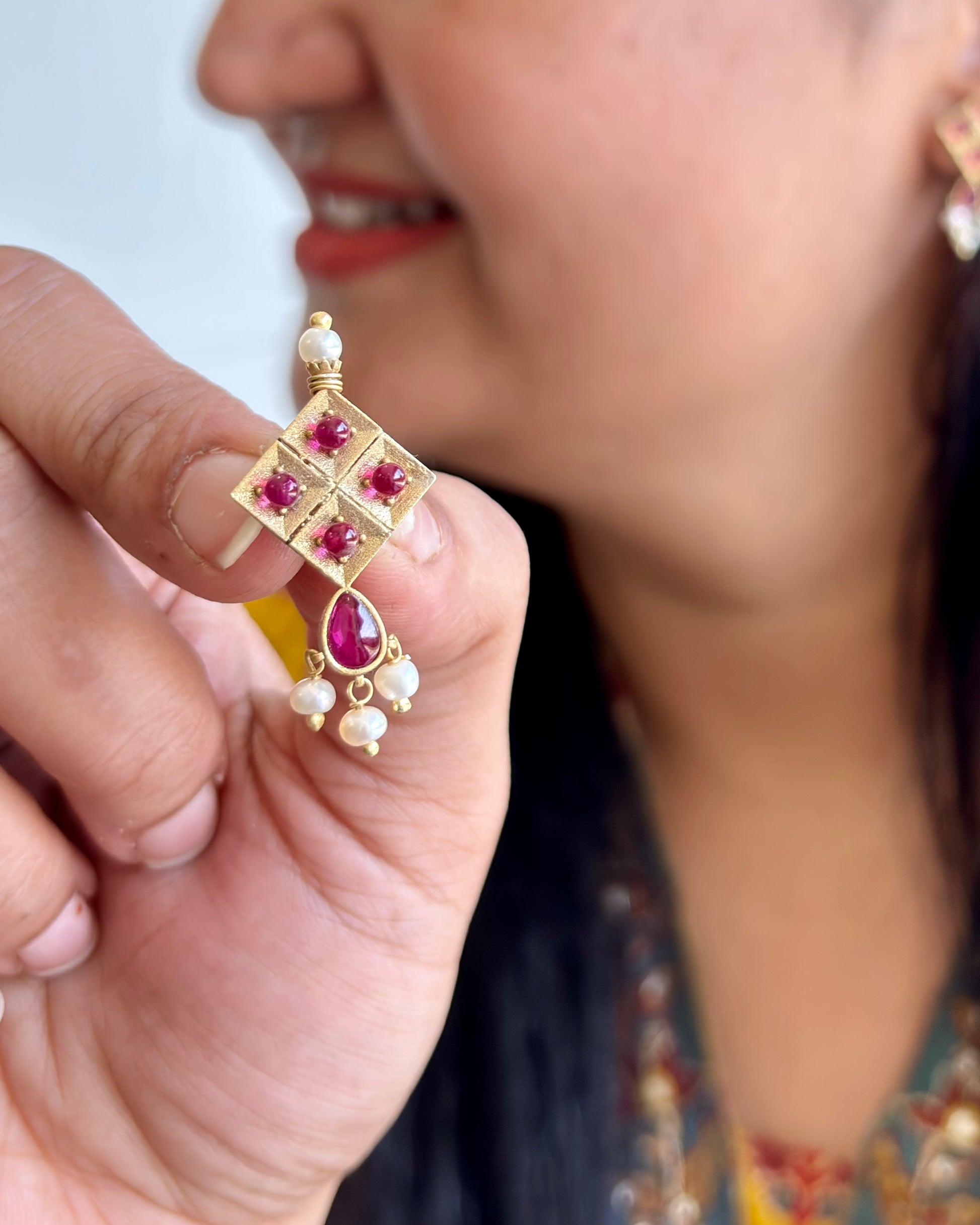 Gold earring with red gemstones held by a hand, blurred background